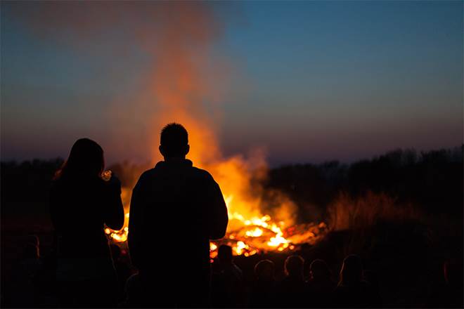 Foto på stor brasa utomhus med två personer i förgrunden