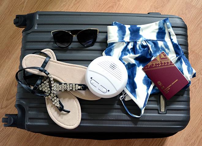 På en resväska ligger badkläder, sandaler, pass och en brandvarnare.